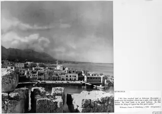 Kyrenia harbour and waterfront seen over ruined stone blocks, with a moored boat and mountains behind the town.