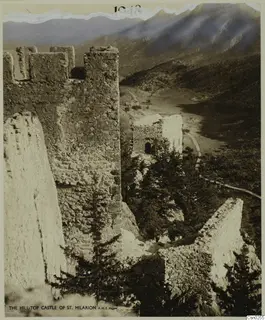 Ruined castle walls with crenellations and a small stone building among pine trees, with mountains behind, on a photo mount
