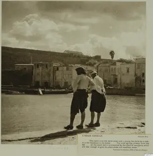 Two men stand with their backs to the camera at the edge of Kyrenia harbour, looking over the water toward houses and a fort wall.