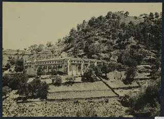 Long stone building with a small tower on a wooded hillside above terraced fields.