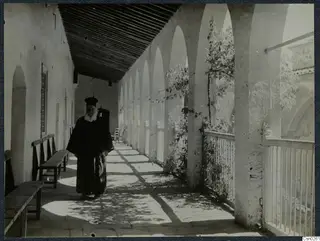 Bearded man in a long dark robe and tall black hat standing in a covered arcade with arches and a tiled floor.