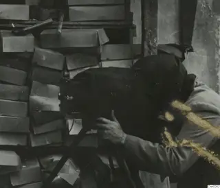 Photographer with head under a dark cloth behind a large camera, with stacked wooden boxes behind.