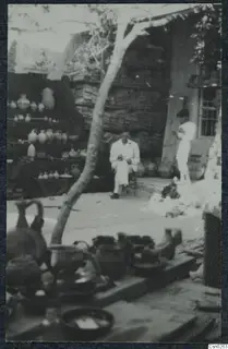 Seated man in a light suit beside shelves of pottery jugs and jars in a courtyard, with a standing figure to the right