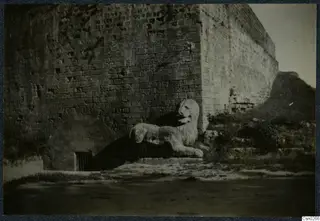 Stone lion sculpture lying beside a large masonry wall and an arched opening with a barred grate
