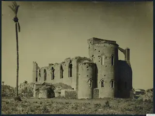 Ruined stone building with narrow arched windows and rounded bays, with a tall palm tree on the left