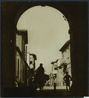 Rider silhouetted under a stone archway, looking down a street of tall buildings with balconies and shutters