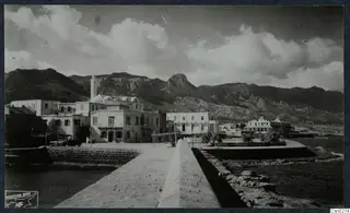 Seafront with a stone quay wall running towards plastered buildings, with rocky mountains and the sea behind and beside them