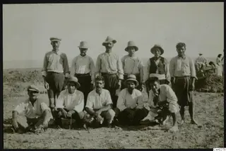 Group of men in hats and caps posing in a dry field, with other workers bending over earth in the background.