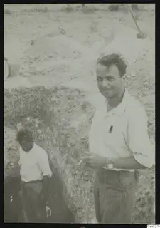 Two men at the edge of an excavation trench, one smiling at the camera, with spoil heaps behind them.