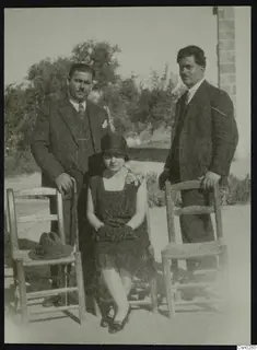 Two men in suits and a seated woman posing with wooden chairs in front of trees