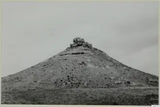 Conical hill with a ruined stone structure on its summit, grassy ground in the foreground under an overcast sky