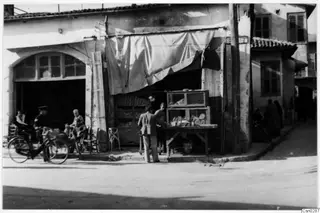 Street corner shop in Nicosia with a cloth awning, a counter and display cases, a bicycle and several people outside