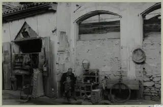 Seated man beside wire cages and a wooden cart outside a plastered building in Nicosia