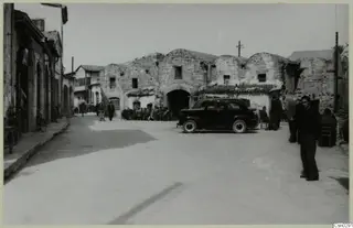 Street in Nicosia with a dark parked car, a stone building with arched openings, and people seated and standing.