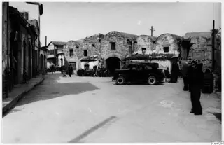 Street in Nicosia with a parked dark car, seated people, and an old stone building with arched gables behind.