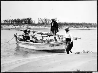 Small wooden boat with lettering on its hull, four men with it in shallow water beside a sandy shore.