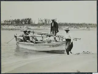 Four people with a wooden boat in shallow water at the edge of a sandy shore, reeds behind them.
