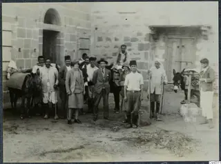 Group of about a dozen people and two donkeys in a dirt yard in front of a stone building with an arched doorway.