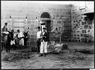 Two young men stand in a dusty courtyard before a stone building, with men and mules waiting at the left.