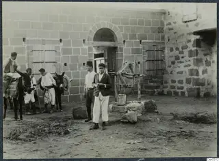 Two young men stand before a stone building with arched doorway, with two mules and three men to the left
