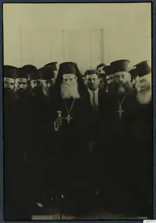 Group of bearded men in black robes and tall black hats, with pectoral crosses; a man in a suit stands among them