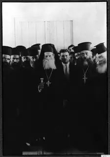Group of bearded men in long black robes and tall black hats, the central man holding a walking stick and a cross on a chain.
