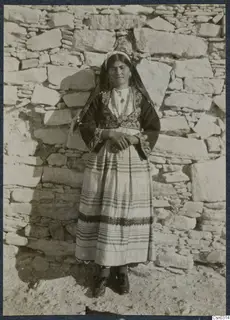 Woman in embroidered traditional dress and headscarf standing before a dry-stone wall