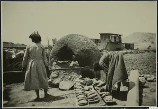 Two women beside a domed clay oven, with flatbreads laid out on boards and a two-storey house behind.