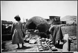 Two women at a domed clay oven, one bending to set out flatbreads on wooden boards, with a two-storey building behind.