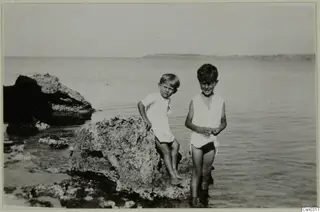 Two boys standing in shallow sea water beside a rough rock, with calm sea behind them
