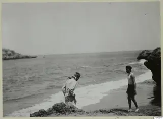 Two men on a sandy beach at the edge of the sea, one bending over by the surf, the other standing near rocks.
