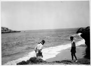 Two men on a sandy beach at the edge of the sea, with rocky headlands along the shore.