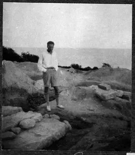 A man in shirt and shorts stands among excavated stone blocks and earth, with the sea behind him.