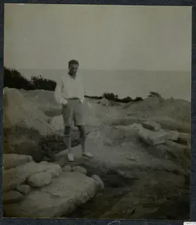 A man in shirt and shorts stands on dug ground beside cut stone blocks, with the sea behind him.