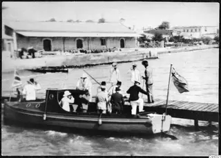 Crown Prince's landing at Limassol: a launch with people aboard approaching a wooden jetty, with flags and a long building on the quay