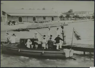 Group of men and a woman in hats on a motor launch alongside a wooden jetty at Limassol
