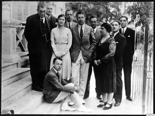 Ten people posing in a group on the stone steps of a house, with an ornate iron gate and a tree at the right.