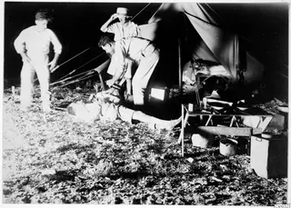 Men at a night camp beside a tent, with a folding table of bowls and pots in the foreground.