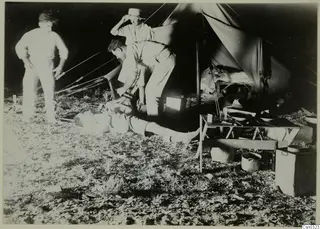 Three men by a canvas tent at night, with a folding table, enamel bowls and cooking pots on the ground