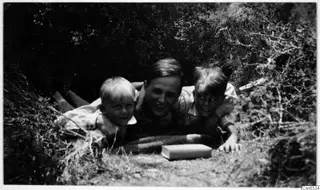 Einar Gjerstad and two children lying side by side in a gap in dense bushes, with a small book on the ground in front of them.
