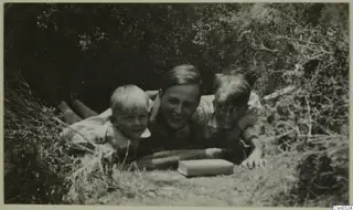 Einar Gjerstad and his two children lying in a gap in bushes, with a book on the ground in front of them