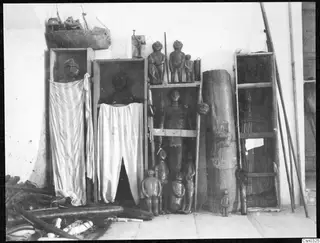 Carved wooden figures and a model ship on wooden shelving, with striped and white cloths hung over two open frames.
