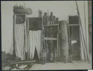 Carved wooden figures and draped cloths on wooden shelves and in open cabinets against a white wall