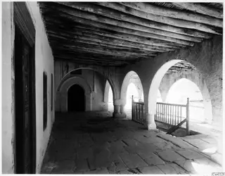Covered arcaded walkway of a monastery building with a timber ceiling, a wooden door and a railing beside an open edge