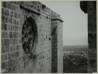 Stone wall with a round tracery window and buttress, with a view over a town and trees below, at Vessberg