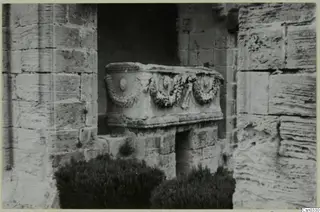Carved stone chest with garland reliefs in a niche of stone blocks, with dark shrubs in front
