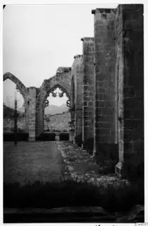 Ruined monastery arcade with pointed arches and a window with stone tracery, seen across a grassy court