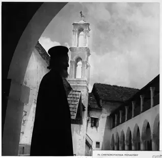 Bearded man in a long dark robe and cap standing under a stone arch, with a bell tower of Chrysorroyiatissa monastery behind him