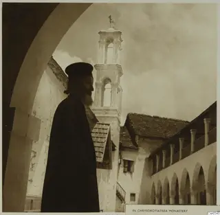 Bearded man in dark robe and black cap in profile under an arch, with the bell tower of Chrysorroyiatissa monastery behind