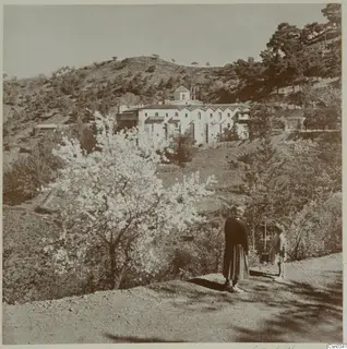 Machairas Monastery across a hillside, with a flowering tree in the foreground and two people on a path